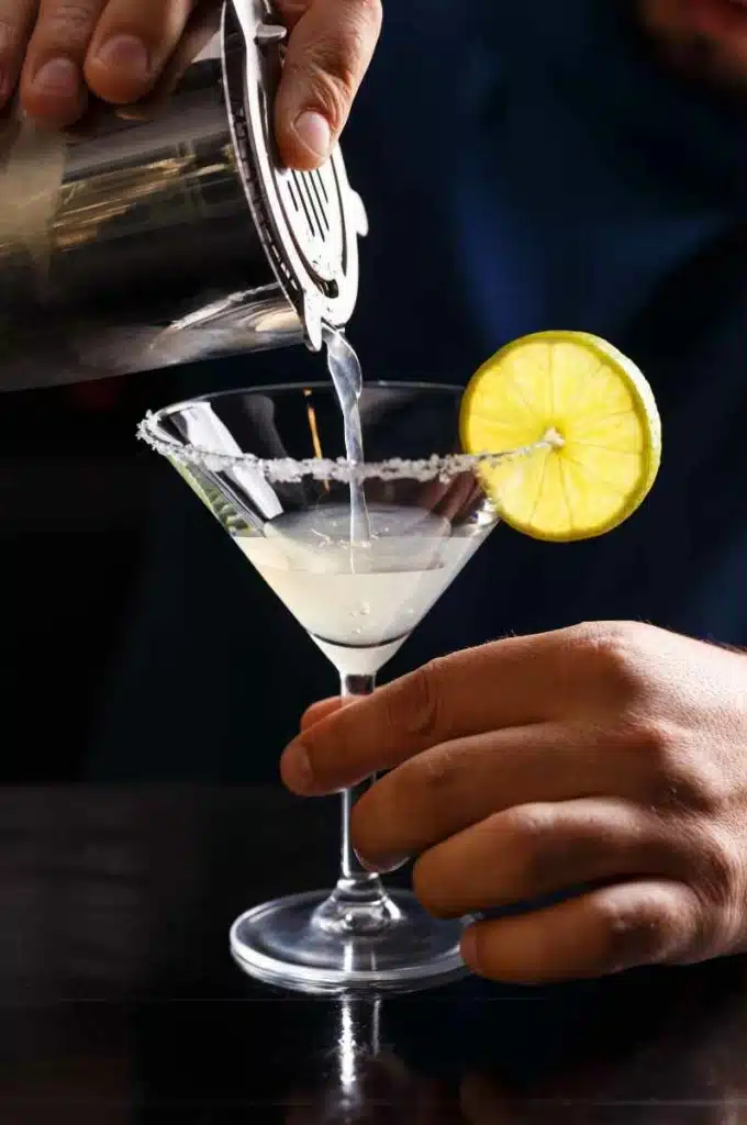 Straining Margarita cocktail into cocktail glass with salt rim and lime wheel